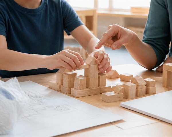 Child rebuilding a wooden block structure while an adult points gently to the next step